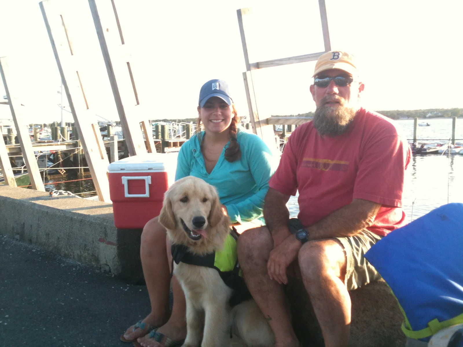 Halcyon Kennels owners Peter and Bridget seated on a Mattapoisett dock with their golden retriever Dory wearing a yellow service vest, southeastern Massachusetts harbor in the background.